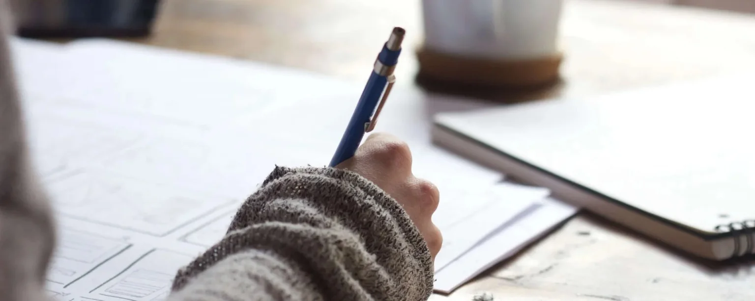 Close-up of a person writing with a blue pen in a notebook on a desk, with papers and a coffee cup in soft focus.