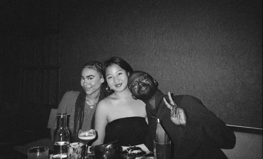 Black-and-white photo of three friends sitting at a restaurant table, smiling at the camera; Joseph Awuah-Darko on the right flashes a peace sign.