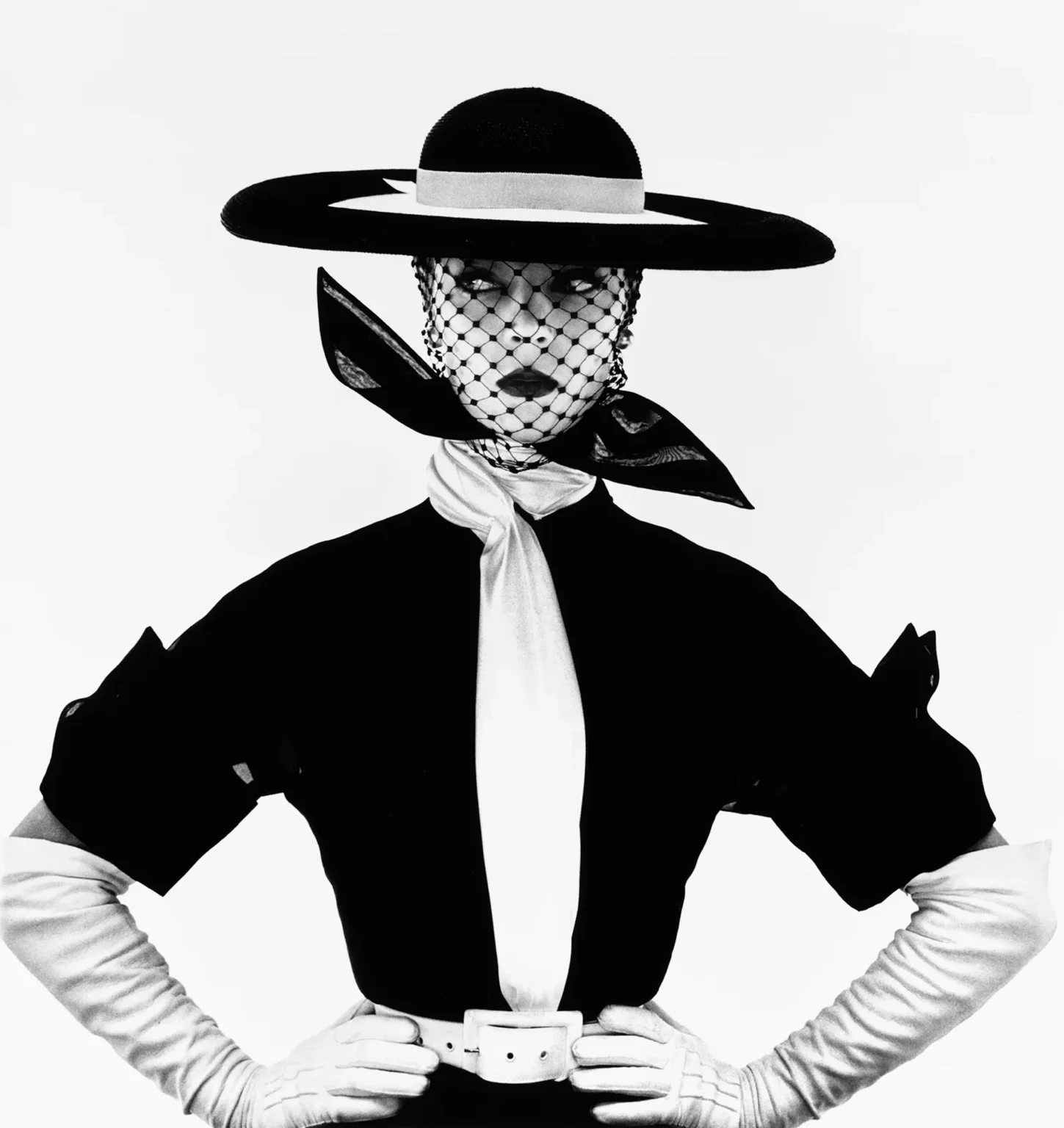 Black-and-white fashion photograph by Irving Penn of a model in a wide-brimmed hat, face veiled in netting, long gloves and a cinched dress, posed against a plain studio backdrop.