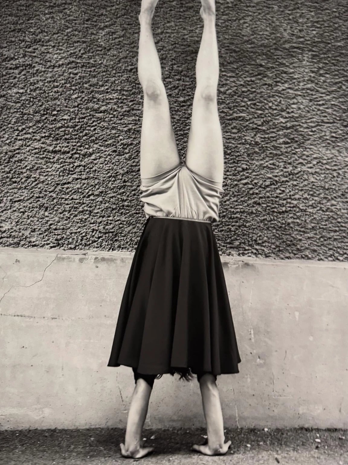 Black-and-white photograph of a person doing a handstand against a rough wall, wearing a knee-length dark skirt and bare feet, with the image framed so the body appears upside down.