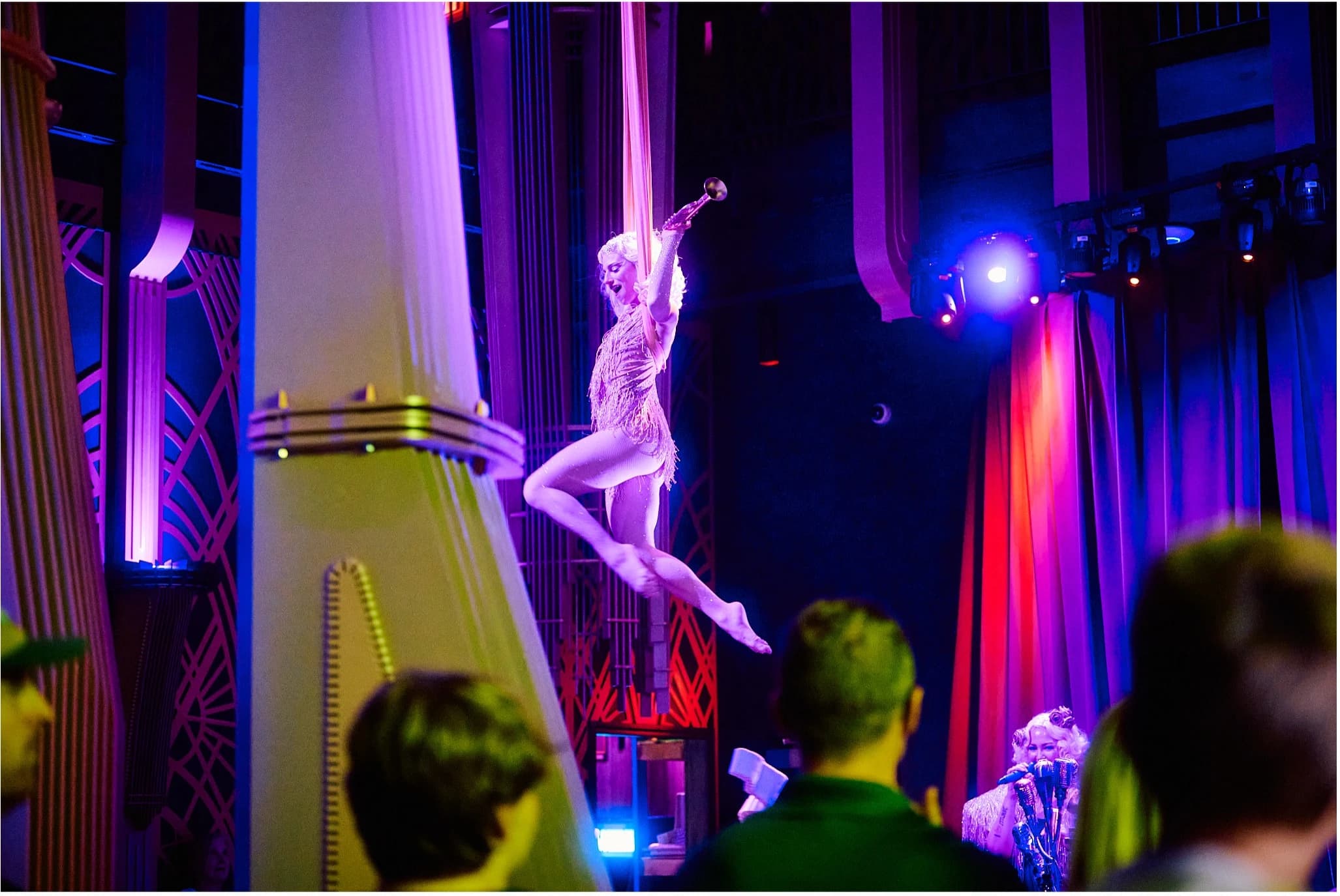 Aerial performer in a fringed costume suspended mid-air above the crowd in a neon-lit jazz club, with dramatic purple and blue stage lighting and audience members watching from below.