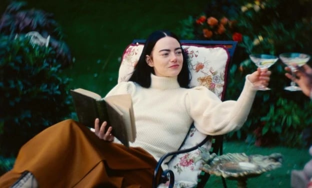 Emma Stones character lounges on a garden chair in Poor Things, holding a book in one hand and raising a cocktail glass in the other, surrounded by lush greenery and flowers.