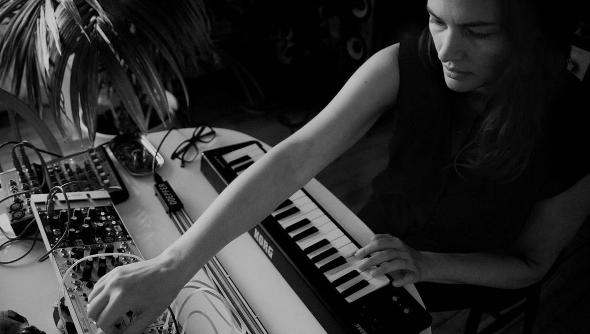 Black-and-white photograph of Christina Vantzou working with a modular synthesizer and a Korg keyboard, her arm reaching across patch cables and controls in an intimate studio setting. Photograph by Julie Calbert.