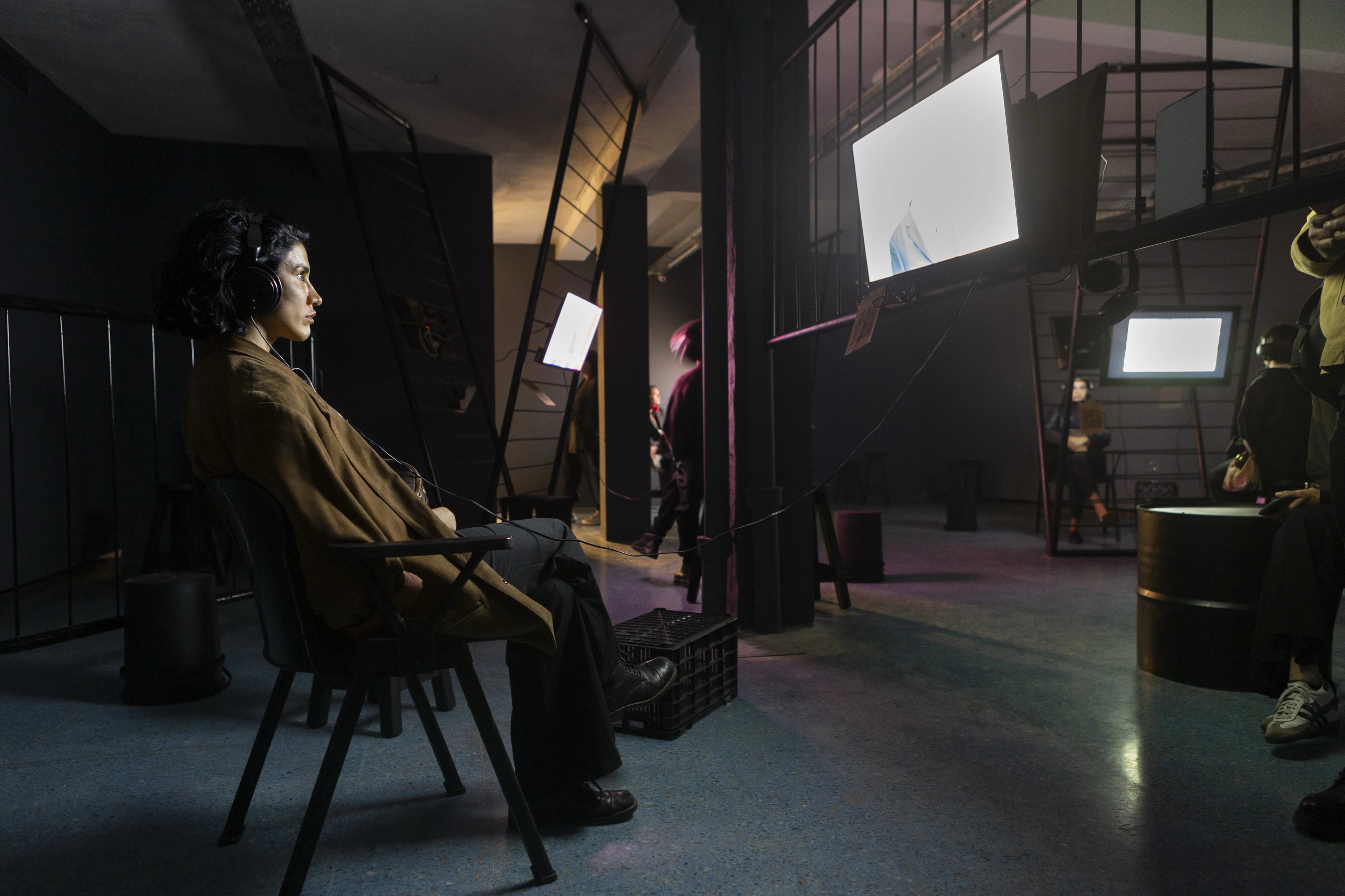 A person wearing headphones sits in a dark studio space, surrounded by bright monitor screens and filming equipment, with crew members in the background.