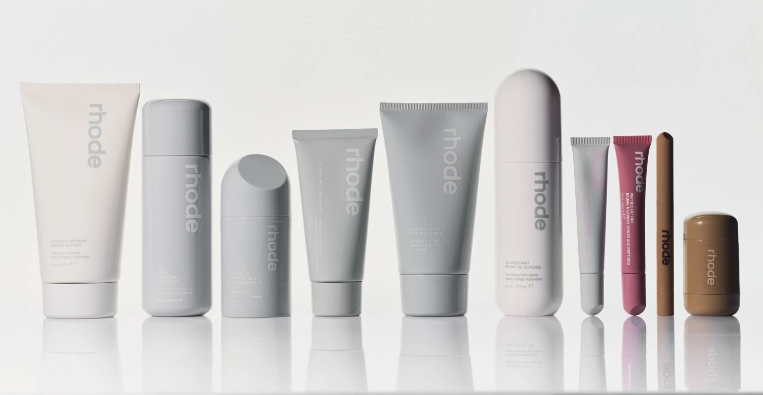 Rhode skincare and lip products arranged in a clean lineup on a light background, featuring minimalist grey, pink, and beige packaging.