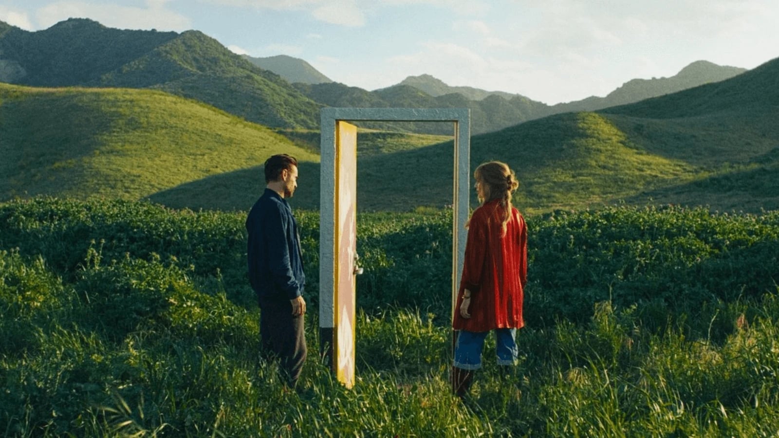 Two characters stand facing a glowing door in a green field with mountains in the background in a still from A Big Bold Beautiful Journey (2025), directed by Kogonada.